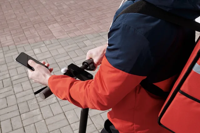 A delivery person riding an electric scooter using a smartphone for navigation outdoors. Reviewing app and publisher delivery reports